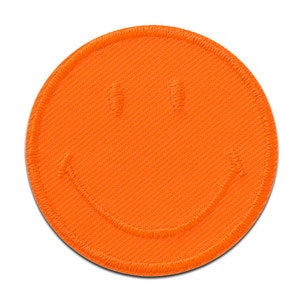 May include: A round, orange patch with a stitched border and a smiling face design. The smiley face has two stitched eyes and a curved stitched mouth. The patch is made of a textured fabric.