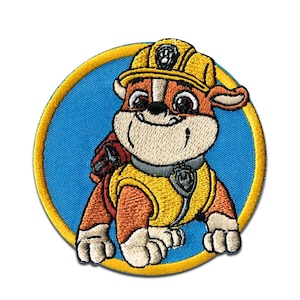 Op de afbeelding: Geborduurde patch met een cartoon bulldog karakter uit de Paw Patrol serie. Het karakter draagt een gele bouwhelm en vest, tegen een blauwe achtergrond met een gele rand. Geschikt voor kleding en accessoires.