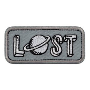 Può includere: Una toppa ricamata grigia con la parola "LOST" in lettere bianche. La "O" è sostituita da un'immagine in bianco e nero di un pianeta con un anello.