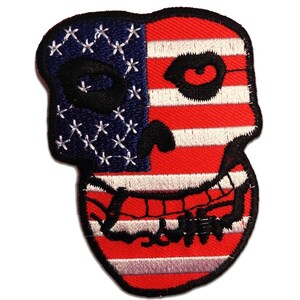 Op de afbeelding: Geborduurde patch in de vorm van een schedel, met het ontwerp van de Amerikaanse vlag. De linkerkant van de schedel is blauw met witte sterren, en de rechterkant heeft rode en witte strepen. De schedel heeft een zwarte omtrek en details.