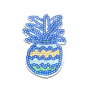 Può includere: Una toppa a forma di ananas blu e gialla fatta di paillettes. L'ananas ha una base verde e una parte superiore blu con una striscia gialla.