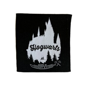 Może przedstawiać: Czarna kwadratowa naszywka z białą sylwetką zamku Hogwart i napisem "Hogwarts" w skrypcie. Poniżej znajduje się napis "School of Witchcraft & Wizardry" ze skrzyżowanymi różdżkami.