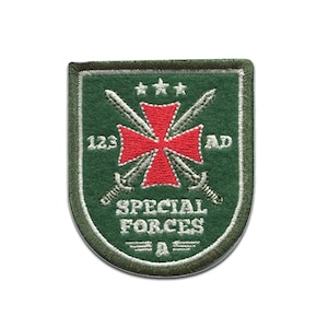 Könnte beinhalten: Gestickter Aufnäher in Form eines grünen Schildes. Der Aufnäher zeigt ein rotes Kreuz, gekreuzte Schwerter und den Text "SPECIAL FORCES" und "123 AD". Der Rand ist grün mit einem genähten Rand.