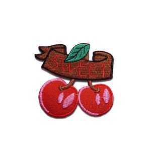 Puede incluir: Parche bordado con dos cerezas rojas con detalles rosas, unidas por tallos marrones. Una pancarta marrón en la parte superior muestra la palabra "SWEET" en rojo, con una hoja verde. El parche está diseñado para ropa o accesorios.