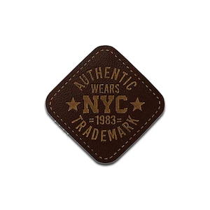 Może przedstawiać: Brązowa skórzana naszywka z napisem "AUTHENTIC WEARS NYC TRADEMARK =1983=". Naszywka ma kształt rombu z zaokrąglonymi rogami i obszytą krawędzią. Dwie gwiazdy flankują napis "NYC".