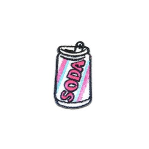 Könnte beinhalten: Gestickter Aufnäher mit einem Limonaden-Dosen-Design. Die Dose ist weiß mit rosa und blauen diagonalen Streifen. Das Wort "SODA" ist rosa, schwarz umrandet. Der Aufnäher hat einen schwarzen Rand.