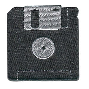 Könnte beinhalten: Ein schwarzer, gestickter Aufnäher in Form einer 8,89 cm großen Diskette. Der Aufnäher hat eine silbernfarbene, verschiebbare Metallabdeckung, eine kreisförmige Nabe und einen rechteckigen Etikettenbereich, die alle mit weißen Nähten umrandet sind. Das Design ist eine nostalgische Anspielung auf die Vintage-Computertechnik.