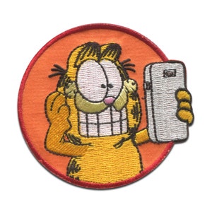 Könnte beinhalten: Ein gestickter Aufnäher von Garfield der Katze, der ein Selfie mit einem weißen Smartphone macht. Der Aufnäher ist orange und schwarz.