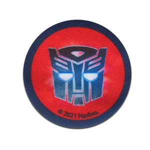 Op de afbeelding: Een ronde patch met het Transformers-logo. Het logo is een gestileerd robotgezicht in blauw en grijs op een rode achtergrond. De patch heeft een marineblauwe rand en de tekst "© 2021 Hasbro".