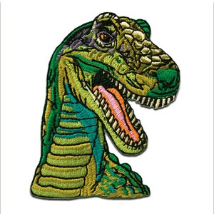 Könnte beinhalten: Grüne, gelbe und blaue gestickte Dinosaurierkopf-Aufnäher. Der Dinosaurier hat einen großen Mund mit scharfen Zähnen und einer rosa Zunge.