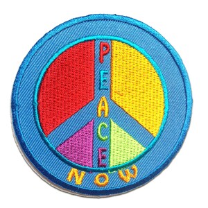 Puede incluir: Un parche azul en forma de símbolo de la paz con la palabra "PEACE" escrita verticalmente en colores arcoíris y la palabra "NOW" debajo.