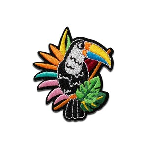 Pode incluir: Patch bordado de um tucano preto com cabeça branca, um bico grande e colorido e uma vibrante variedade de folhas e flores tropicais. O patch tem uma borda preta e é projetado para aplicação em roupas ou acessórios.