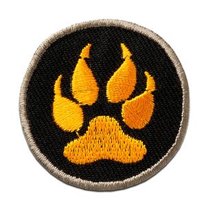 Op de afbeelding: Ronde geborduurde patch met een gouden pootafdruk op een zwarte achtergrond. De pootafdruk is gedetailleerd gestikt en de patch heeft een beige rand. Geschikt voor kleding of accessoires.