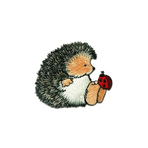 Könnte beinhalten: Gestickter Aufnäher mit einem niedlichen Igel mit einem stacheligen, schwarz-braunen Rücken, einem beigen Gesicht und Pfoten sowie einem weißen Bauch. Der Igel hält einen roten Marienkäfer mit schwarzen Punkten. Auf weißem Hintergrund.