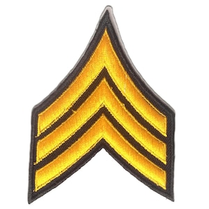 Op de afbeelding: Een goud en zwart geborduurde patch met drie chevrons, gerangschikt in een opwaartse pijl. De patch heeft een zwarte rand en is waarschijnlijk een militair ranginsigne.
