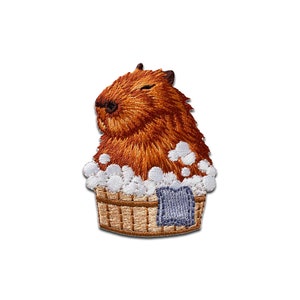 May include: Embroidered patch showing a relaxed capybara enjoying a bath. The capybara is brown with a textured appearance, sitting in a woven tub filled with white bubbles. A small blue towel hangs on the tub's side. The patch has a white border.