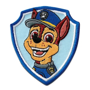 Op de afbeelding: Geborduurde patch met Chase van Paw Patrol. De schildvormige patch heeft een lichtblauwe achtergrond en een blauwe rand. Chase, een Duitse herdershond-puppy, wordt afgebeeld met een blauwe politiepet en uniform, glimlachend.