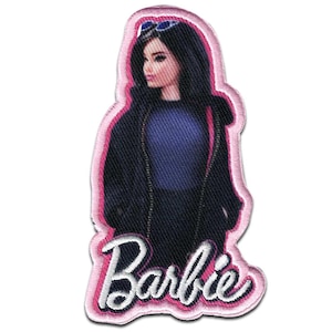 Op de afbeelding: Een geborduurde patch met een pop met lang zwart haar en een blauwe zonnebril bovenop haar hoofd. De pop draagt een donkerblauw topje onder een zwart jasje. Het woord Barbie is onderaan in wit schrift geborduurd met een roze rand.