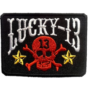 Könnte beinhalten: Schwarzer gestickter Patch mit weißem Text "LUCKY-13" und einem roten Totenkopf mit Kreuzknochen und zwei gelben Sternen.