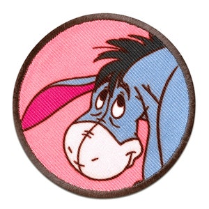 Könnte beinhalten: Ein runder, rosa und brauner gestickter Aufnäher mit einem Cartoon-Bild von Eeyore, dem Esel aus Winnie Puuh. Eeyore schaut mit einem traurigen Gesichtsausdruck nach unten.