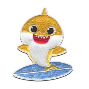 Könnte beinhalten: Ein gestickter Aufnäher mit einem gelb-weißen Cartoon-Hai mit großen schwarzen Augen und einem roten Mund, der auf einem hellblauen Surfbrett steht. Das Design hat einen weißen Rand und ist in einem flachen, gestickten Textilstil gehalten.
