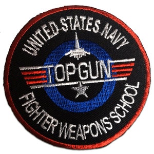 Może przedstawiać: Okrągła, haftowana naszywka z napisem "UNITED STATES NAVY TOPGUN FIGHTER WEAPONS SCHOOL". Naszywka ma czarne tło, niebieskie koło oraz czerwone i białe akcenty. Zawiera samolot odrzutowy i gwiazdę.