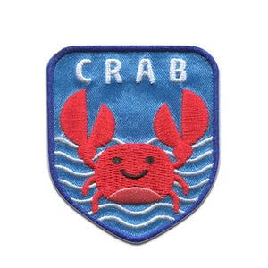 Peut inclure: Écusson bleu en forme de bouclier avec un motif de crabe rouge. Le crabe a un visage souriant et se détache sur un fond bleu avec des lignes de vagues blanches. Le mot "CRAB" est brodé en blanc.