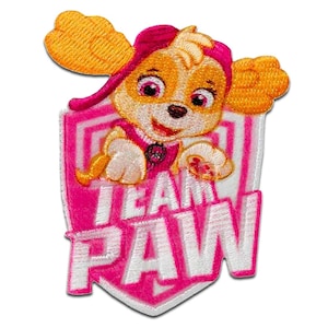 Könnte beinhalten: Gestickter Aufnäher mit Skye von Paw Patrol. Der Aufnäher zeigt Skye, einen hellbraunen Welpen mit rosa Akzenten, in einem rosa-weißen Schild. Die Worte "TEAM PAW" sind in Weiß auf das Schild gestickt.