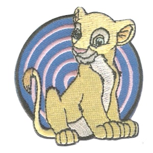 Op de afbeelding: Geborduurde patch met een cartoon leeuwenwelp in tinten geel, beige en wit. De leeuwenwelp staat tegen een blauwe cirkelvormige achtergrond met roze en witte concentrische lijnen. De patch is bedoeld voor kleding of accessoires.