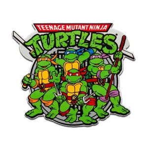 Può includere: Una toppa con le Tartarughe Ninja. Le tartarughe verdi sono raffigurate con le loro maschere e armi colorate. Il testo "TEENAGE MUTANT NINJA TURTLES" è visualizzato sopra le tartarughe in rosso e verde.
