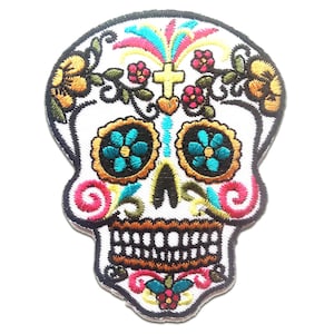 Puede incluir: Parche bordado de calavera con fondo blanco, con diseños florales y de cruz coloridos. Los ojos de la calavera son flores turquesas con centros negros, y la boca tiene una hilera de dientes. El parche está delineado en negro.