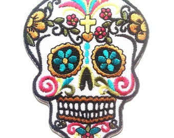 May include: Embroidered skull patch with a white background, featuring colorful floral and cross designs. The skull's eyes are turquoise flowers with black centers, and the mouth has a row of teeth. The patch is outlined in black.