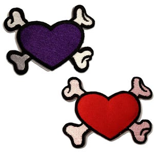 Puede incluir: Dos parches bordados con un diseño de corazón con huesos cruzados. Un parche tiene un corazón morado con detalles en blanco y negro, mientras que el otro tiene un corazón rojo con detalles similares. Los parches están sobre un fondo blanco.