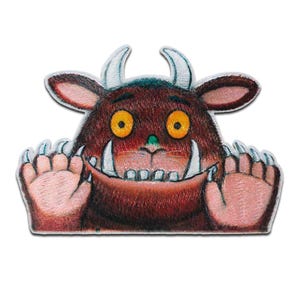 Op de afbeelding: Een bruin monster met grote gele ogen en scherpe tanden kijkt over een muur. Het monster heeft twee hoorns en glimlacht.
