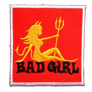 Può includere: Toppa ricamata con sfondo rosso e bordo bianco. La toppa presenta una donna diavolo gialla che tiene un tridente, con le parole "BAD GIRL" in nero.