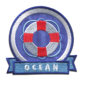 Op de afbeelding: Geborduurde patch met een blauw en wit reddingsboei-ontwerp met rode accenten. De patch heeft een blauwe banner aan de onderkant met het woord "OCEAN" in witte letters. De patch is cirkelvormig en heeft een witte rand.