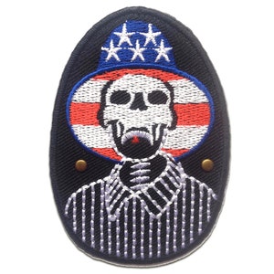 Peut inclure: Un écusson brodé de forme ovale sur fond noir. L'écusson représente un crâne blanc avec un nez rouge et une moustache noire. Le crâne porte un chapeau bleu et blanc avec cinq étoiles blanches. Le chapeau est posé sur un cercle rouge, blanc et bleu sur fond blanc. Le cercle a une bande rouge en haut et en bas et une bande blanche au milieu. L'écusson a deux boutons dorés sur les côtés.