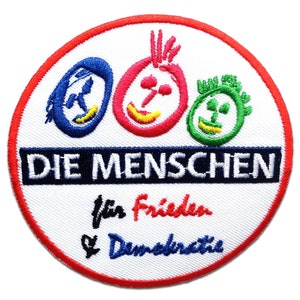 Peut inclure: Écusson brodé circulaire avec un fond blanc et un bord rouge. Il présente le texte allemand "DIE MENSCHEN" en bleu marine, avec trois visages de dessins animés au-dessus. En dessous, les mots "für Frieden & Demokratie".