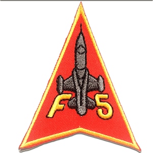 Peut inclure: Écusson brodé avec un avion à réaction gris sur un fond triangulaire rouge. L'écusson porte le texte "F5" en jaune, souligné de noir, et une bordure jaune.