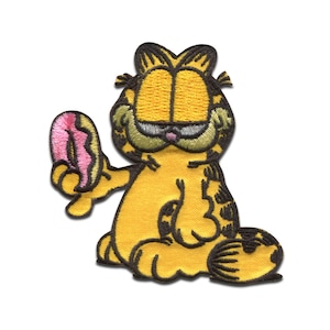 Puede incluir: Un parche bordado para planchar con Garfield el gato sosteniendo una rosquilla rosa. El parche es amarillo, naranja y negro.