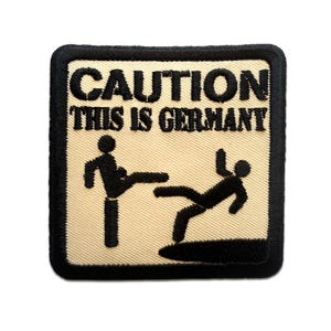 Könnte beinhalten: Gestickter Aufnäher mit dem Text "CAUTION THIS IS GERMANY" über einer Strichmännchenfigur, die eine andere Strichmännchenfigur tritt. Der Aufnäher hat einen beigefarbenen Hintergrund mit schwarzem Text und einem schwarzen Rand.