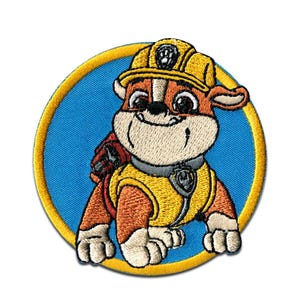 May include: Embroidered patch featuring a cartoon bulldog character from the Paw Patrol series. The character is wearing a yellow construction hat and vest, set against a blue circular background with a gold border. The patch is designed for application on clothing or accessories.