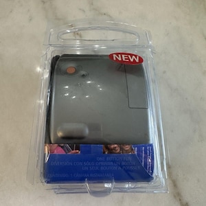 Peut inclure: Un appareil photo instantané argenté dans un emballage plastique transparent, avec un autocollant rouge "NEW". L'emballage comprend du texte en plusieurs langues, dont "ONE BUTTON FUN" et "1 CAMARA INSTANTANEA".