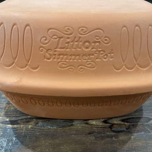 May include: A terracotta-colored Litton Simmer Pot. The pot has a wide, shallow base and a domed lid. The word "Litton Simmer Pot" is embossed on the side of the pot. The pot has decorative oval and dot patterns around the rim.