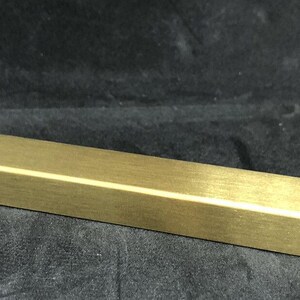 Pode incluir: Um puxador de armário retangular dourado com um design em forma de L em cada extremidade. A ferragem tem um acabamento escovado e foi projetada para fácil instalação. O puxador tem aproximadamente 15 cm de comprimento.