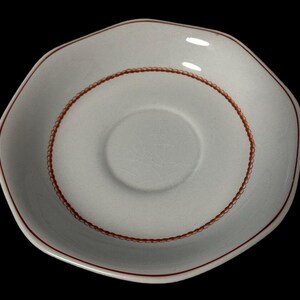 Peut inclure: Une soucoupe en céramique blanche de forme irrégulière. Le bord et un anneau décoratif central sont soulignés en brun rougeâtre. La soucoupe possède un petit centre en retrait, adapté à une tasse à thé ou un petit mug.