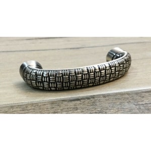 May include: A silver cabinet pull with a woven pattern. The pull is curved and has a smooth, polished finish.