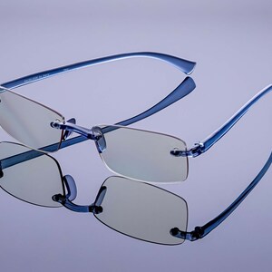 Puede incluir: Gafas sin montura azules con un marco azul claro y lentes transparentes. Las gafas est&aacute;n sobre una superficie de color p&uacute;rpura claro.