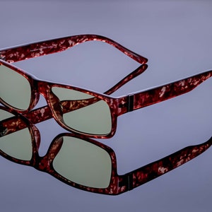 Könnte beinhalten: Eine Brille mit roten Schildpattbügeln, braunem Gestell und grünen Gläsern. Die Brille ist zusammengeklappt und liegt auf einer reflektierenden Oberfläche.