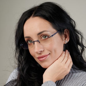 Puede incluir: Una mujer con el pelo largo y negro que lleva gafas sin montura con monturas azules. Lleva un jersey gris y tiene la mano en la barbilla.